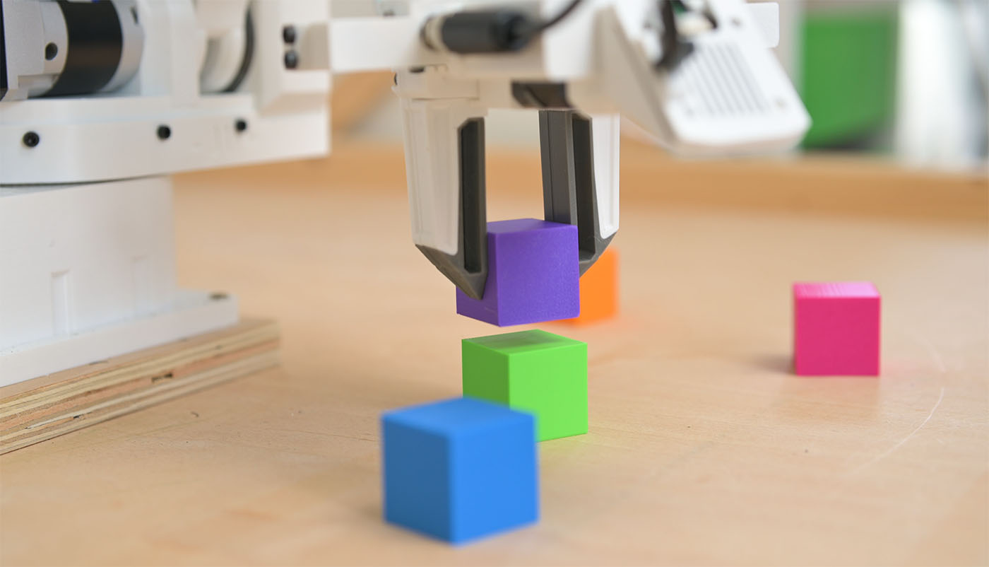 A robot gripper holding a purple block above a green one on the bench